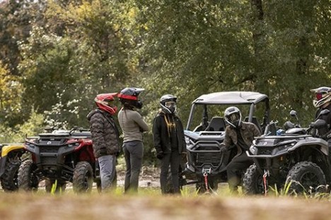 People with helmets stand near all-terrain vehicles in a wooded area.