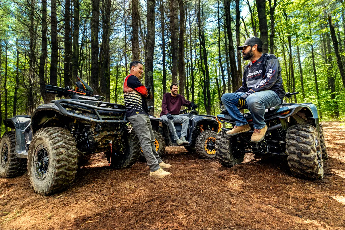 Three men with ATVs chat in a forest with tall trees and sunlight filtering through.