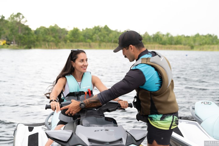A woman on a jet ski with a man assisting her near a lake, both wearing life vests.