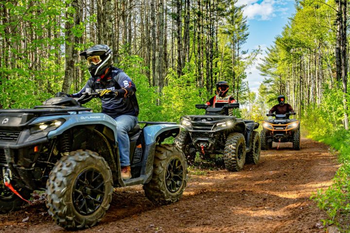Three people riding ATVs on a forest trail surrounded by green trees.