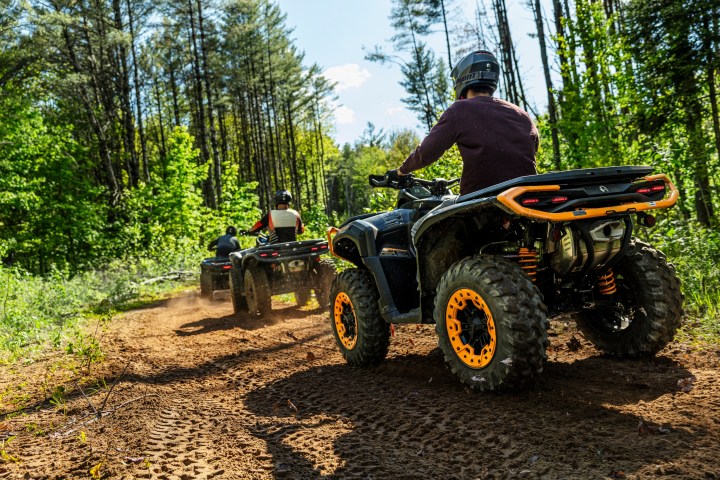 Two people riding ATVs on a forest trail with trees and sunlight.