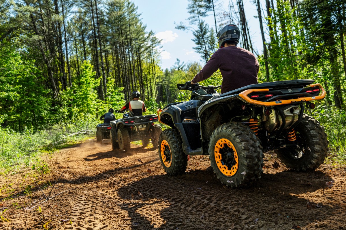 Two people riding ATVs on a forest trail with trees and sunlight.