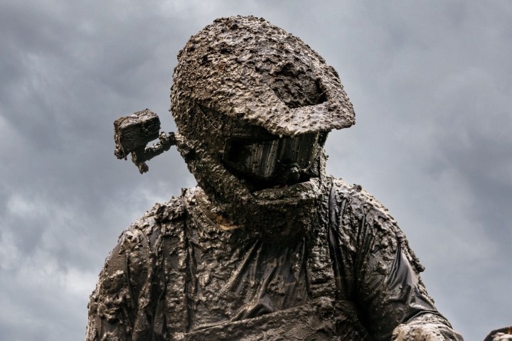 Person covered in mud, wearing a helmet, with cloudy sky in background.