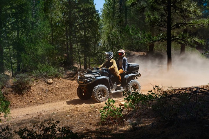 Two people riding an ATV on a dirt trail in a forest, raising dust behind them.