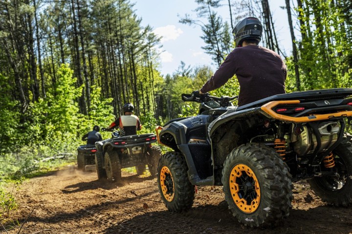 Two people riding ATVs on a forest trail surrounded by trees on a sunny day.