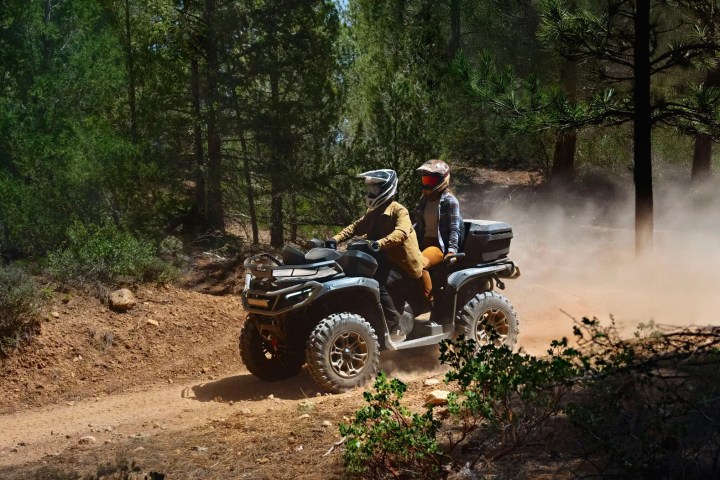 Two people riding an ATV on a dusty forest trail with trees surrounding them.