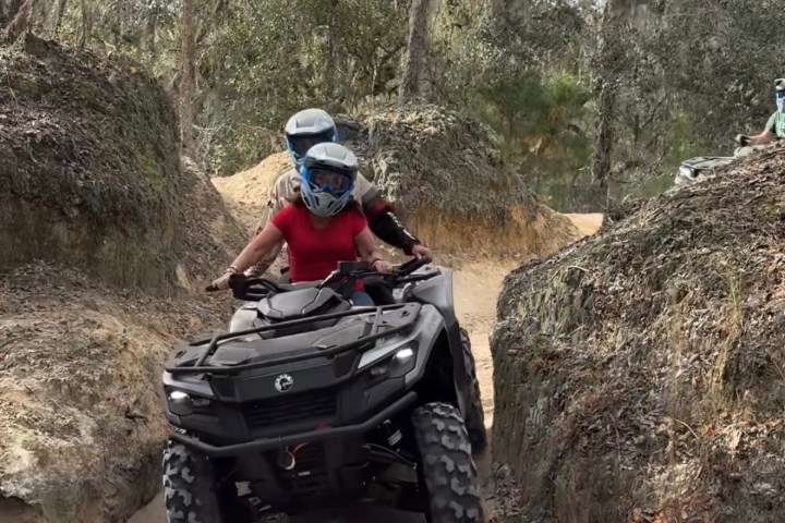 Two people riding an ATV through a narrow dirt path with trees around them.