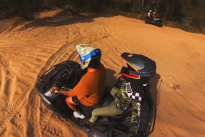 Two people riding ATVs on a sandy trail with trees in the background.