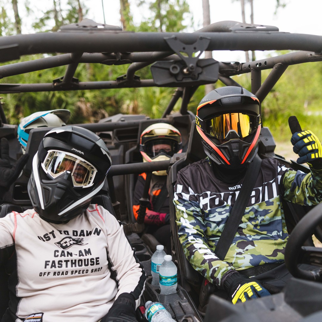 Four helmeted individuals in an off-road vehicle, two gesturing with thumbs up.