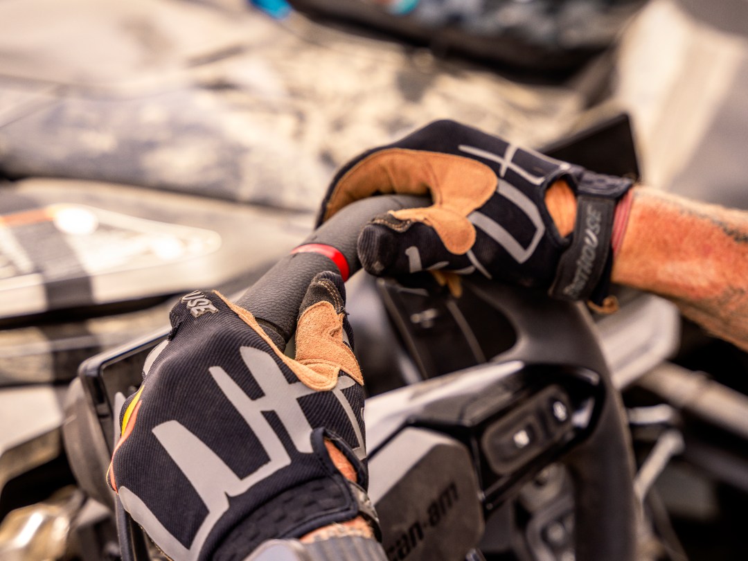 Gloved hands steering an off-road vehicle, with a helmet visible in the background.