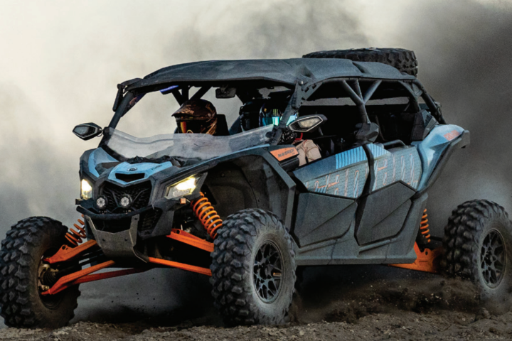 Off-road vehicle kicking up dust on a dirt path, with two occupants wearing helmets.