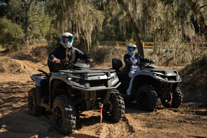 Two people riding ATVs on a dirt trail surrounded by trees and foliage.