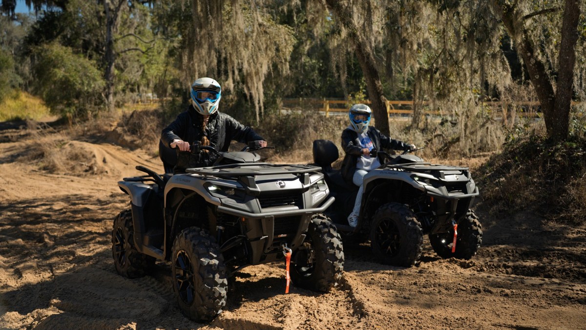 Two people riding ATVs on a dirt trail surrounded by trees and foliage.