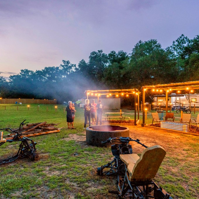 People gather around a fire pit with string lights and seating at dusk in a grassy area.