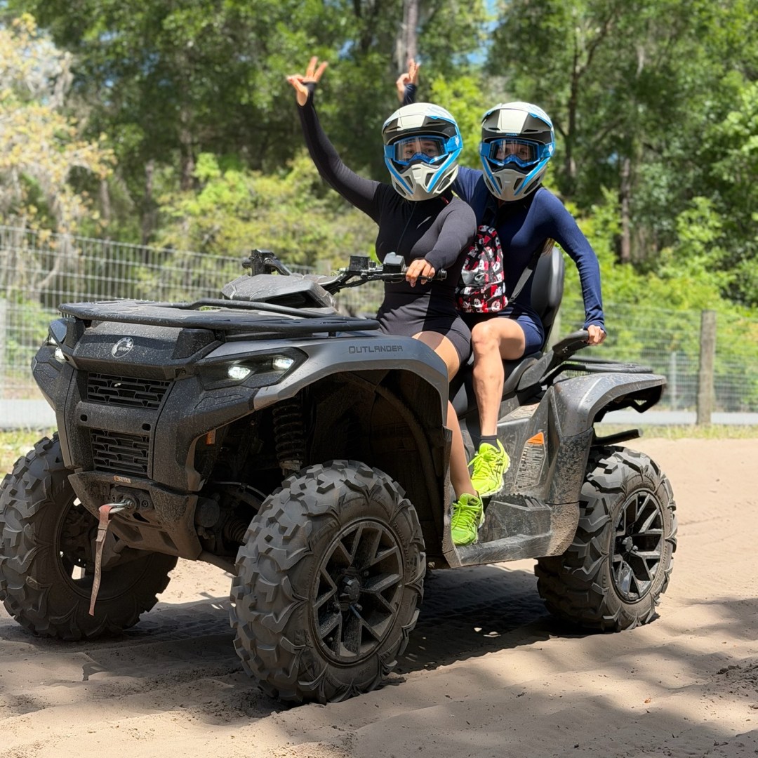 Two people wearing helmets riding an ATV on a dirt path with trees in the background.