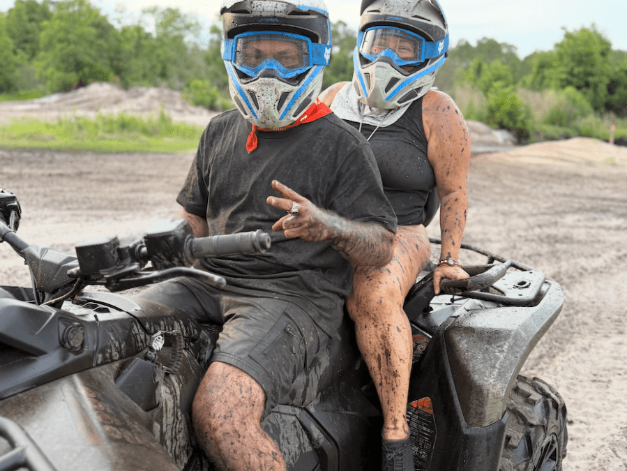 Two people in helmets covered in mud riding an ATV outdoors.