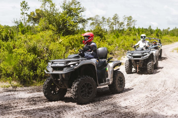 Two people riding ATVs on a dirt path in a forested area.