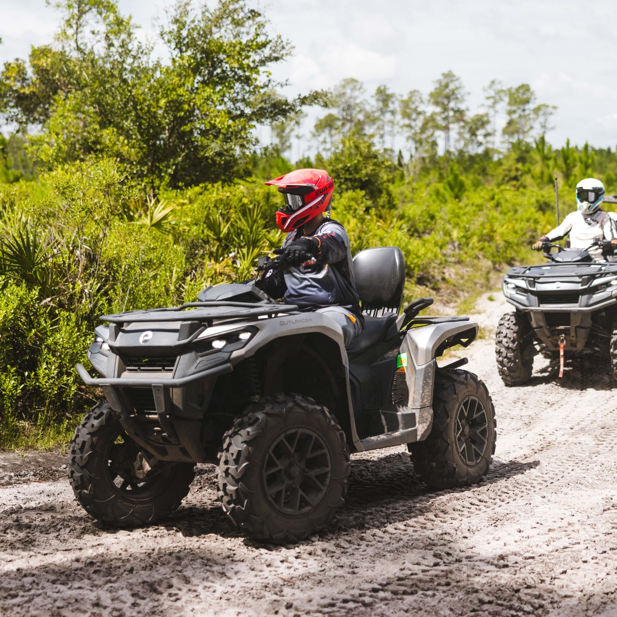 Two people riding ATVs on a dirt path in a forested area.